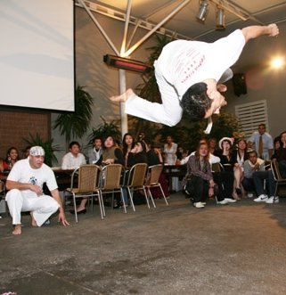Instrutor Futa doing an acrobatic jump while a crowd watches in the background.