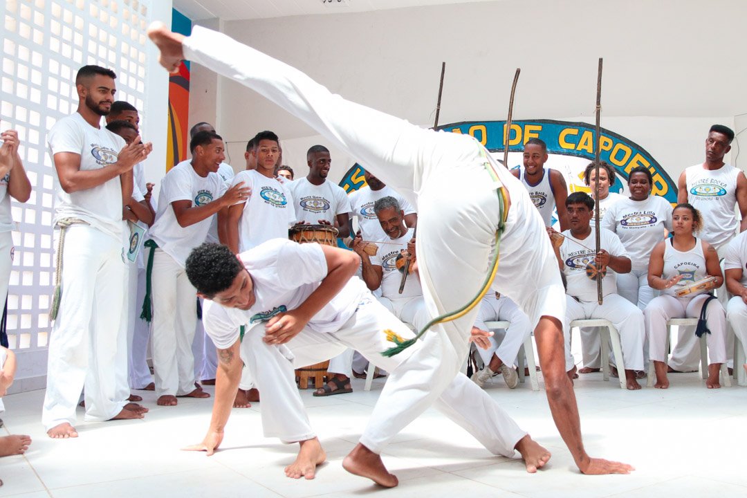 A group of people in white pants and white shirts emblazoned with the Capoeira Angonal logo gather around two people in the foreground, one crouching to dodge the other person's attack, a kick with one hand touching the ground.